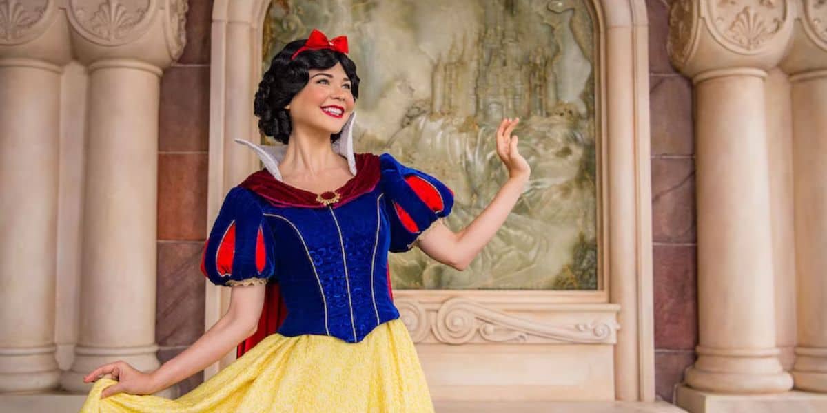 A person dressed as a fairytale character stands in front of a decorative backdrop featuring a castle at Disney World Princess Week. They wear a red bow, blue and red top, and yellow skirt, posing with one hand raised. The setting is ornate with stonework.