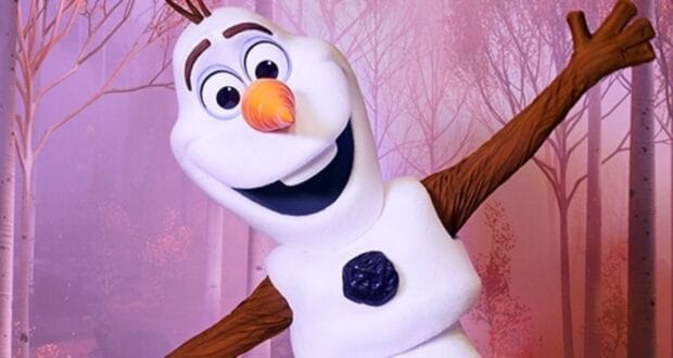 A cheerful snowman with a carrot nose, expressive eyes, and stick arms stands joyfully against a backdrop of pinkish trees in the enchanting world of Disneyland Paris's Frozen Land. His mouth is open in a wide smile, capturing the magic of this icy wonderland.
