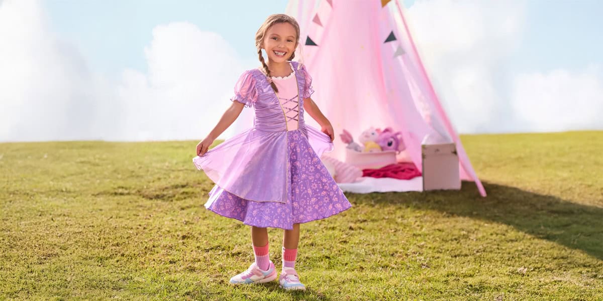 A young girl in a purple princess dress smiles and holds her skirt while standing on grass, celebrating Disney Princesses at home with a pink play tent and toys in the background on a sunny day.