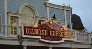A two-story building with a light blue facade and white trim features an illuminated sign that reads "The Diamond Horseshoe" in decorative gold lettering on a red background. The sign is adorned with a horseshoe and several outdoor lights, marking one of the best places to eat at Magic Kingdom.