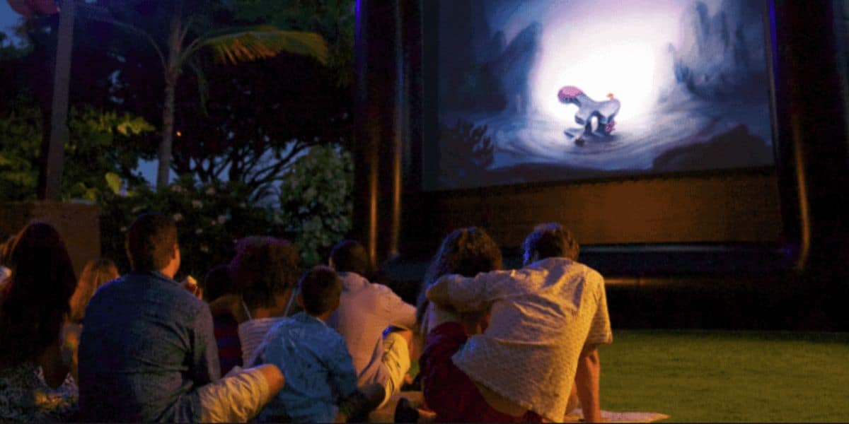 A group of people sit on the grass at night, watching an animated movie on an inflatable outdoor screen. The screen displays a scene with colorful animated characters. Trees and dim lights are visible in the background, reminiscent of magical nights enjoying freebies at Disney World.