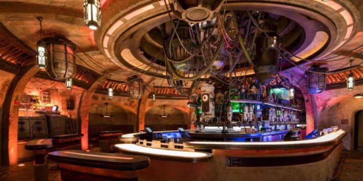 Futuristic bar with a circular design, illuminated by colored lights. The ceiling boasts a large, intricate apparatus with wires and lights above the counter area. Known for serving some of the best drinks at Disney World, it exudes an industrial, science fiction aesthetic.