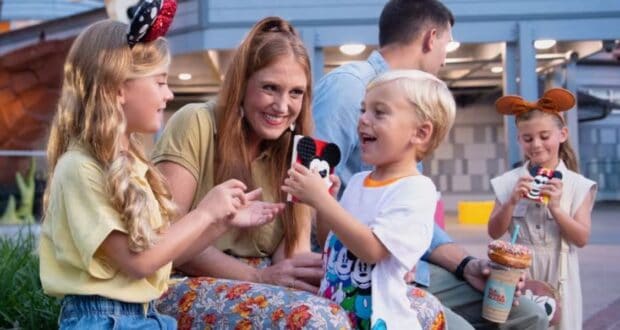A family of Disney Annual Passholders with two children in Mickey Mouse-themed clothing sits outside. The children are smiling, clutching Disney treats, while an adult watches them affectionately. In the background, another child holds a large donut and drink.