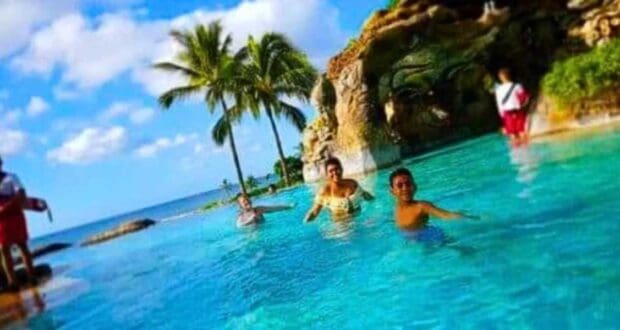 People enjoying swimming in a clear blue pool near the ocean at Disney Aulani. There are palm trees and a rocky structure with greenery in the background under a partly cloudy sky.