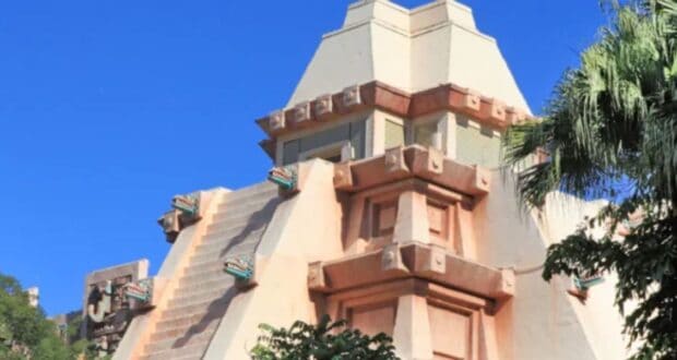 A beige pyramid-shaped structure with a terraced design rises against a bright blue sky, reminiscent of EPCOT's iconic architecture. The building features decorative elements and green accents. In the foreground, lush green trees partially obscure the view, inviting you to savor an afternoon of leisure and drinking.