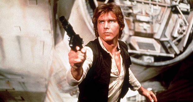 Han Solo in his signature vest takes aim with his blaster inside Star Wars: Galaxy’s Edge, with the Millennium Falcon visible behind him.