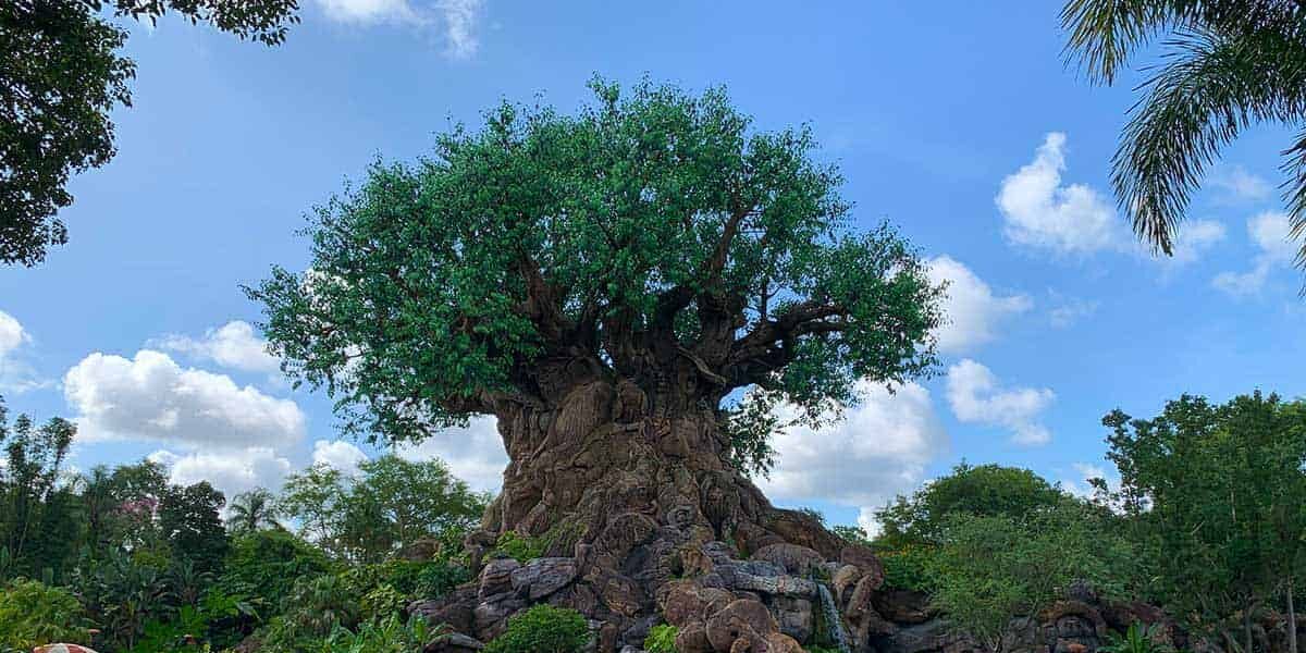 A large, intricately-carved tree stands in lush greenery under a blue sky with scattered clouds. The tree’s trunk features detailed animal sculptures, and leafy green branches extend outward, creating a broad canopy.