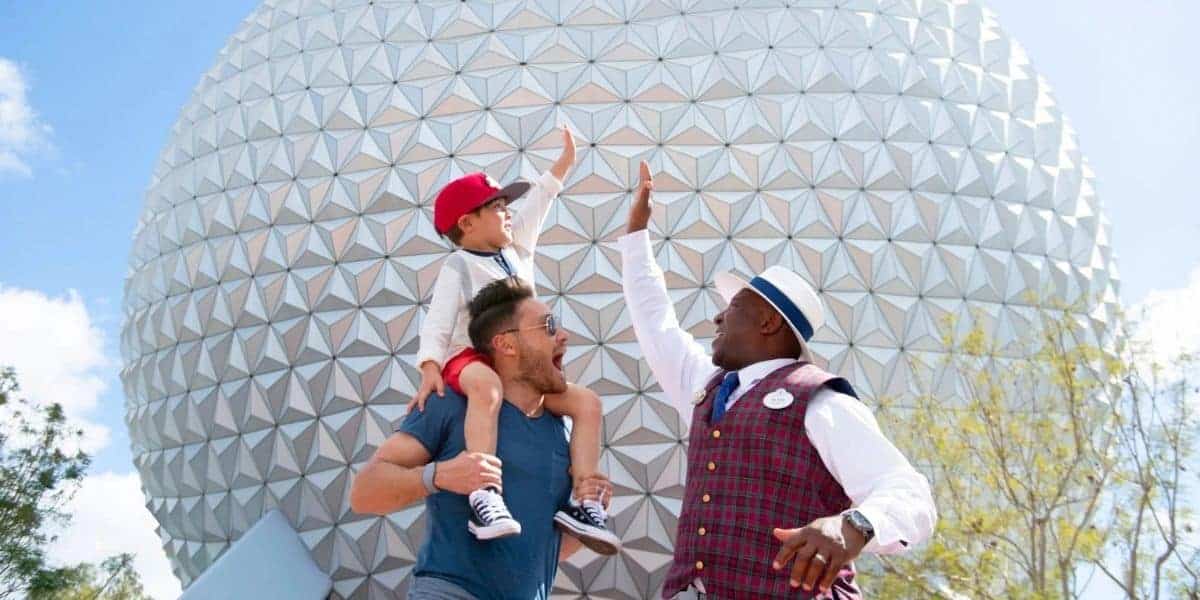 A child riding on an adult’s shoulders shares a high-five with a Disney Cast Member in front of Epcot’s iconic Spaceship Earth, showcasing the joy and magic of experiencing Disney backstage moments.