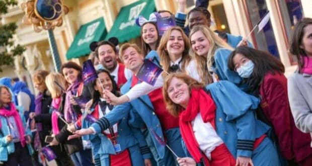 A group of smiling people in blue uniforms and red scarves wave small flags while standing in a crowded outdoor setting, resembling a festive parade or celebration. Some wear mouse ears, and one person wears a mask.