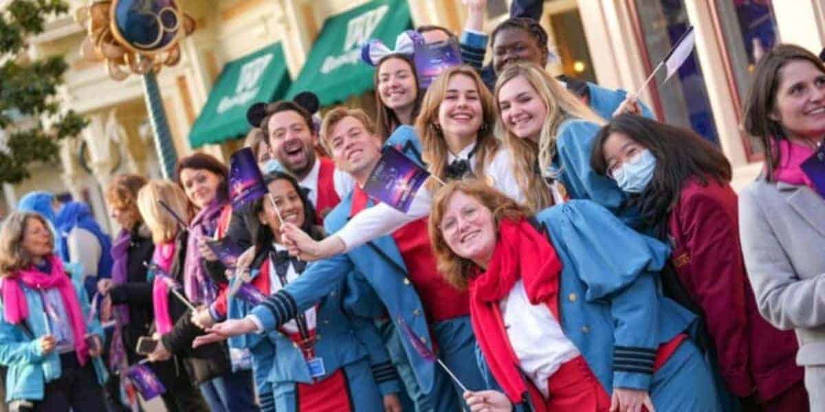 A group of smiling people in blue uniforms and red scarves wave small flags while standing in a crowded outdoor setting, resembling a festive parade or celebration. Some wear mouse ears, and one person wears a mask.