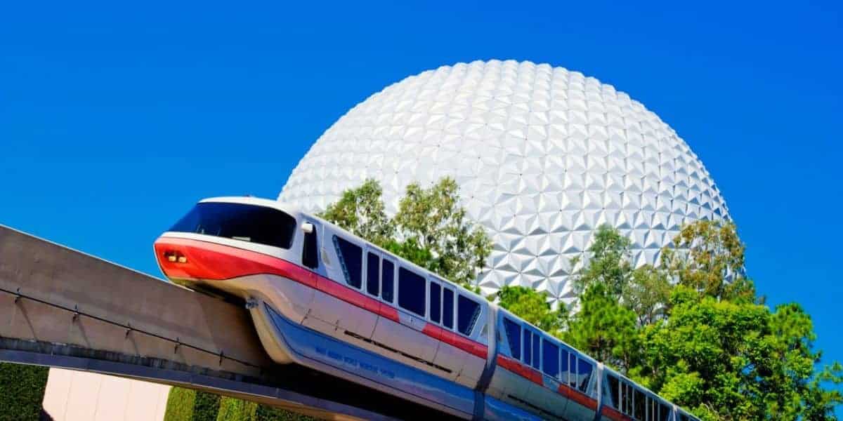 The iconic Walt Disney World Monorail glides past lush trees with Spaceship Earth’s massive geodesic dome rising in the background, all beneath a perfect blue sky at EPCOT.