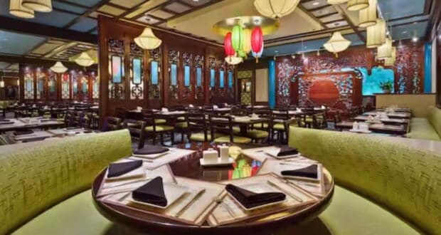 Immersive Asian-themed dining space with circular tables, vibrant green booths, glowing lanterns, and intricate wooden accents, offering guests a transporting experience reminiscent of a theme park's exotic restaurant attraction.
