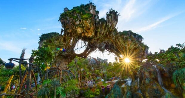 Fantasy landscape with floating mountains covered in lush greenery and vines, under a clear blue sky with sunlight streaming through. Exotic plants and pathways are visible below, creating an otherworldly atmosphere.
