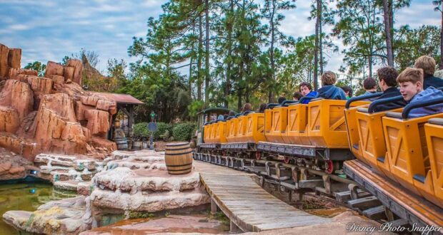 Guests experience the excitement of Big Thunder Mountain Railroad as the yellow train races through rugged, western-inspired canyons surrounded by lush trees beneath a clear blue sky.