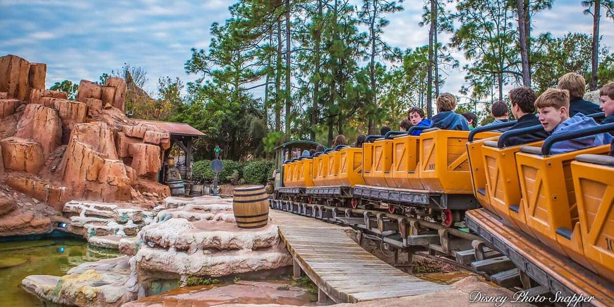 Guests experience the excitement of Big Thunder Mountain Railroad as the yellow train races through rugged, western-inspired canyons surrounded by lush trees beneath a clear blue sky.