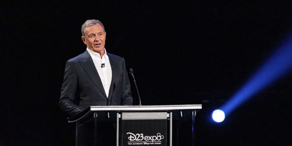 A man in a dark suit speaks at a podium with a microphone, with the "D23 Expo" logo on the front, against a dark background with a bright spotlight.