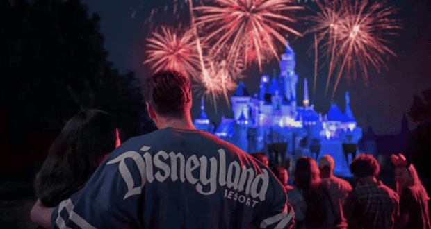 A couple stands in front of a crowd watching a vibrant fireworks display over a brightly lit castle at night. The person in focus is wearing a "Disneyland Resort" shirt.