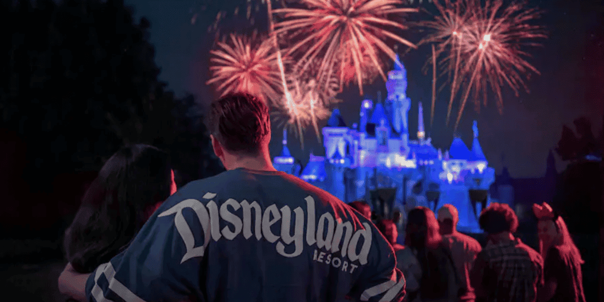 A couple stands in front of a crowd watching a vibrant fireworks display over a brightly lit castle at night. The person in focus is wearing a "Disneyland Resort" shirt.