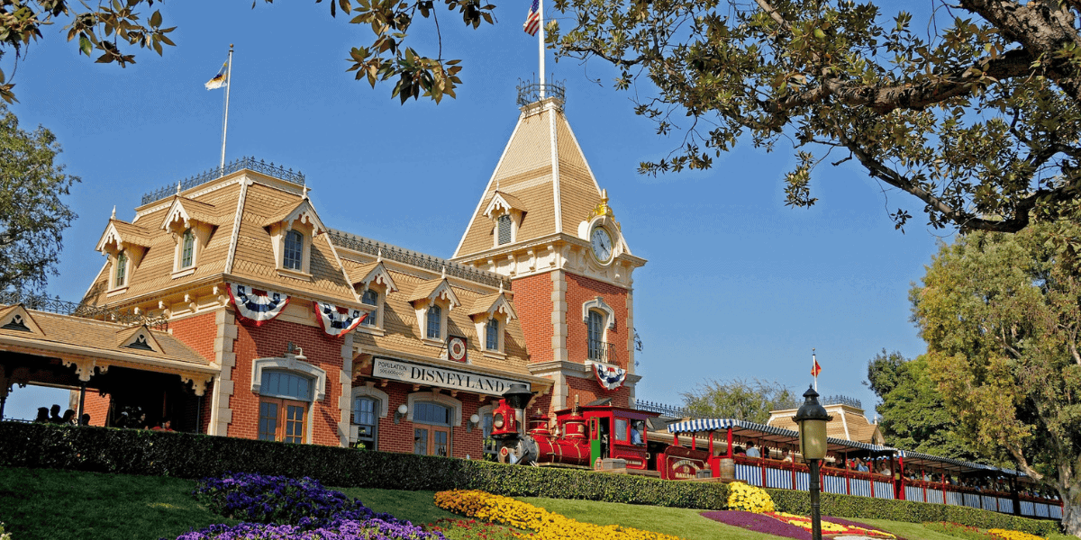 A vibrant vintage train is stationed in front of a large, red-brick building adorned with flags and banners. The foreground features a lush garden with colorful flowers. A clear blue sky and trees surround the scene.