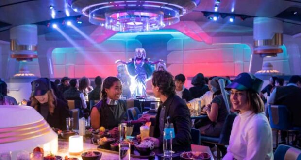 People dine in a futuristic, space-themed restaurant with colorful lighting. A performer in an elaborate alien costume entertains guests at the front of the room. Tables are filled with food and drinks.