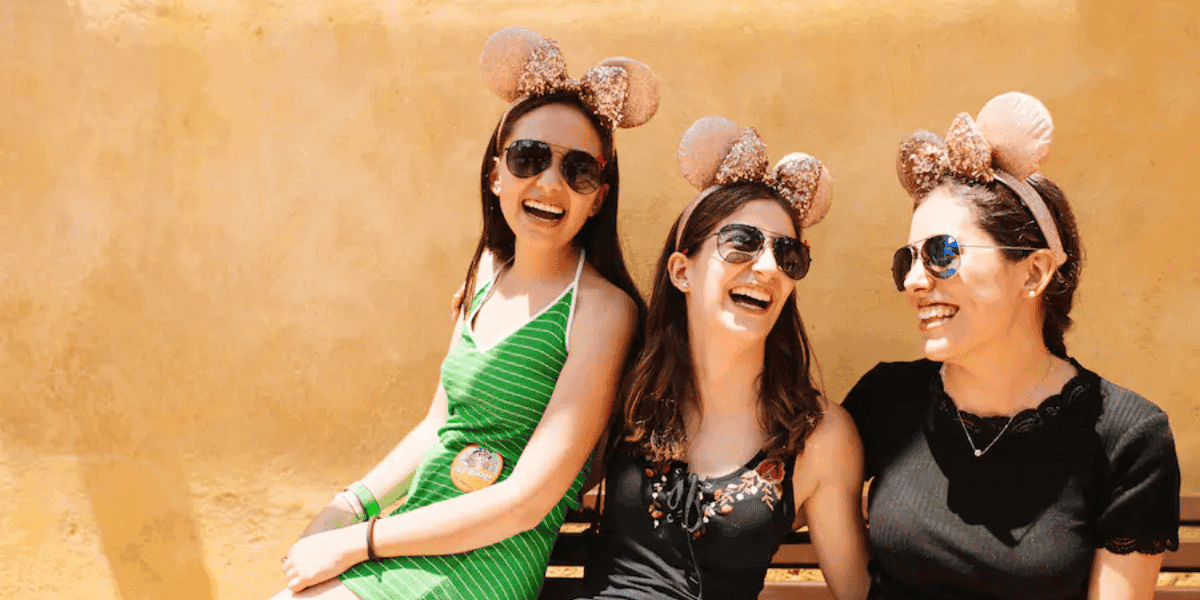 Three women wearing Disney-inspired ear headbands and sunglasses sit on a bench, laughing together against a yellow wall. The woman on the left is in a green dress, the middle in a black top, and the right in a dark dress—bringing cheer even during any Disneyland blackout.