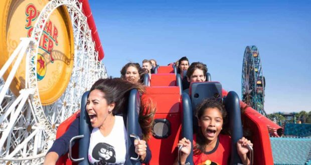 People of various ages scream and smile while riding a red roller coaster at Pixar Pier in an amusement park, with a large Pixar Pier sign and Ferris wheel visible in the background under a clear blue sky.