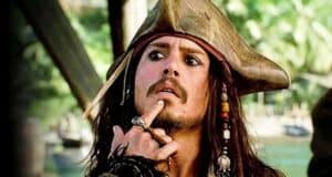 A person dressed as a pirate with a tricorn hat, long hair, beads, and rings touches their chin thoughtfully, channeling a Johnny Depp vibe. Lush greenery and wooden structures appear in the blurred background.