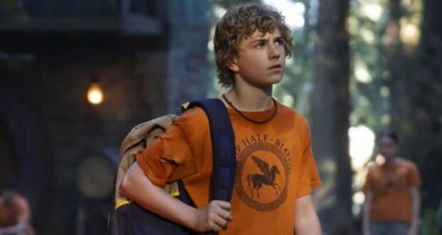 Percy Jackson, with his signature curly hair and orange Camp Half-Blood shirt, stands in a lush, wooded setting at the theme park’s immersive mythological adventure area, gazing out as if ready to embark on his next epic quest.