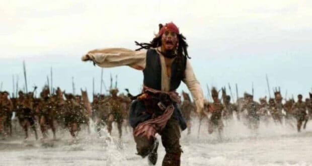 A man dressed as a pirate runs frantically through shallow water, chased by a large group holding spears in the background—a scene reminiscent of fans eagerly awaiting news on the Pirates Caribbean 6 release date.