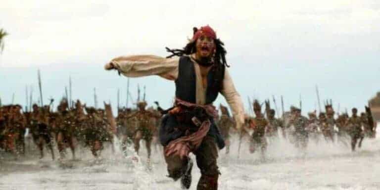 A man dressed as a pirate runs frantically through shallow water while a large group of people wielding spears chase him—a scene reminiscent of waiting for the Pirates Caribbean 6 release date.