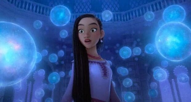 An animated heroine marvels as glowing blue orbs float around her in the magical, grand hall of a fantasy-themed attraction.