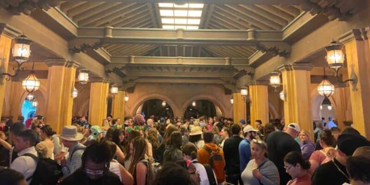 A large crowd gathers under a covered walkway adorned with wooden beams and lanterns. The bustling atmosphere is filled with excitement as people wait, some clutching their phones accessing Disney Genie and wearing hats. Numerous arches create a magical backdrop in the distance.