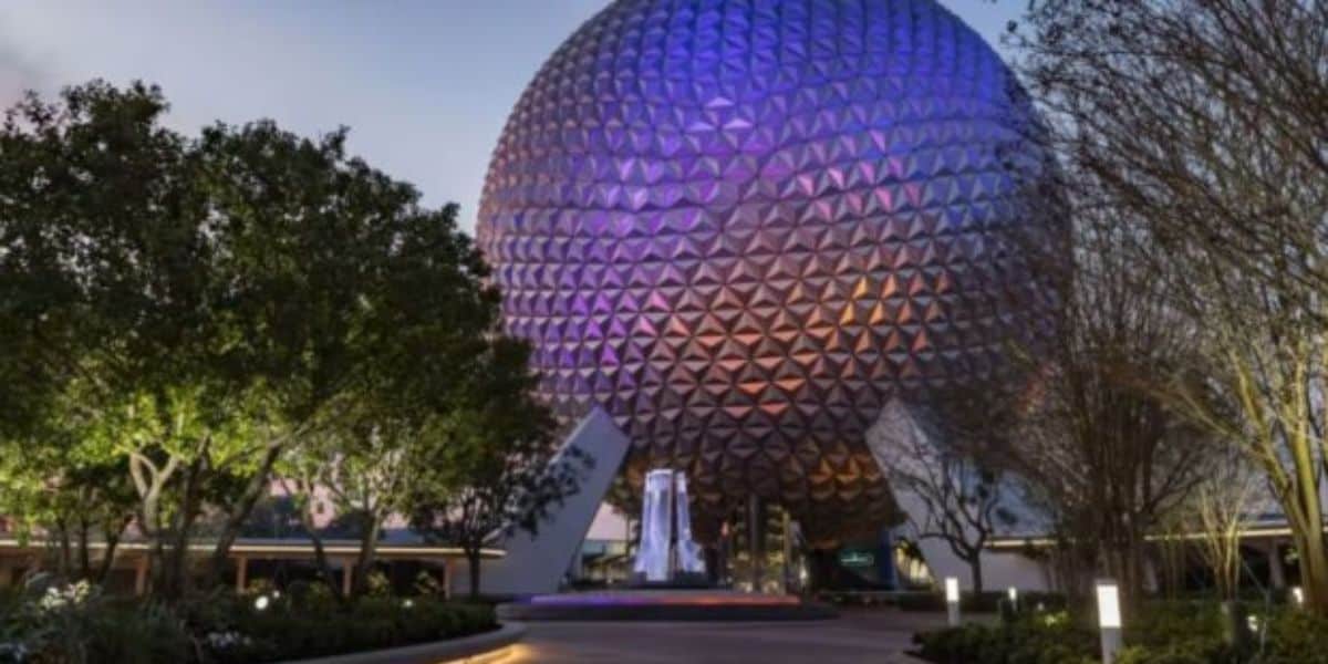 A geodesic sphere with purple lighting stands regally at Disney World, surrounded by trees and pathways. A small fountain dances in front, under a clear sky.