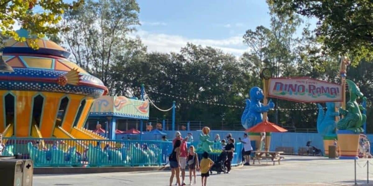 People stroll through a vibrant section of Animal Kingdom, featuring a spaceship-themed ride on the left and dinosaur decorations holding a "Dino-Rama!" sign on the right. In the background, trees frame the scene against a clear sky.