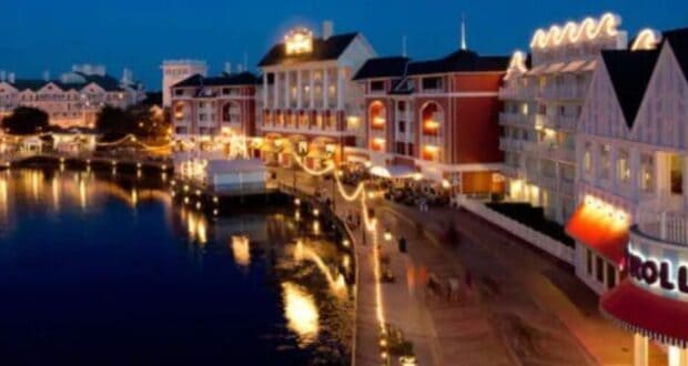 A vibrant waterfront scene at night features brightly lit buildings reflected in the water. The Disney World Boardwalk, lined with decorative lights and a couple of shops, including one with a red and white awning near Jellyrolls, adds to the lively atmosphere.