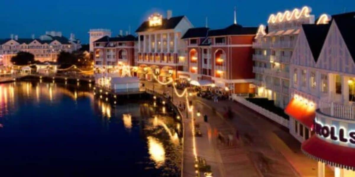 A vibrant waterfront scene at night features brightly lit buildings reflected in the water. The Disney World Boardwalk, lined with decorative lights and a couple of shops, including one with a red and white awning near Jellyrolls, adds to the lively atmosphere.