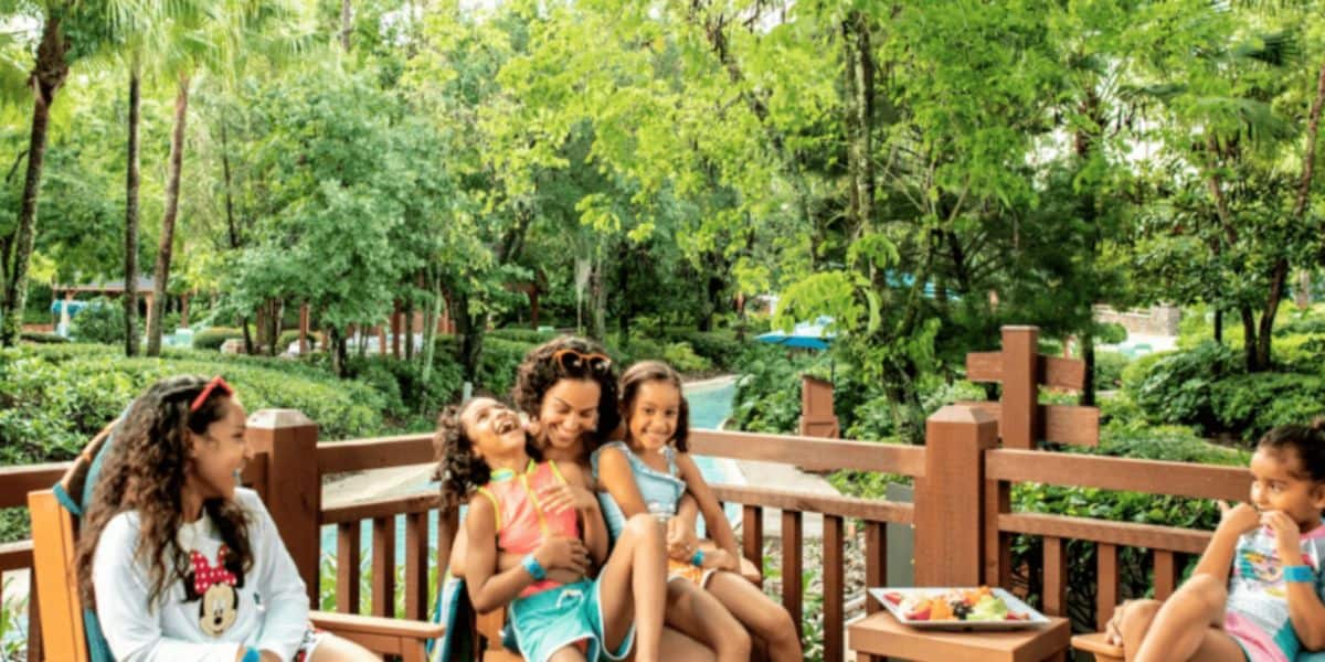 A group of children and a woman are sitting on a wooden deck surrounded by lush greenery, feeling the enchantment of a Disney Theme Park. They enjoy a relaxed moment together with smiles and laughter, while a table with snacks adds to the magic nearby.