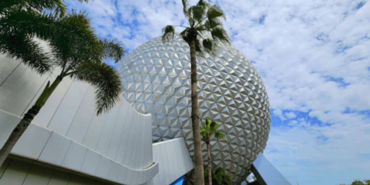 A large, geodesic sphere with a silver, triangular panel exterior stands timelessly against a partly cloudy sky. Palm trees line the foreground while a white, modern building curves gracefully on the left—a serene scene undisturbed by any tragedy at Disney World.