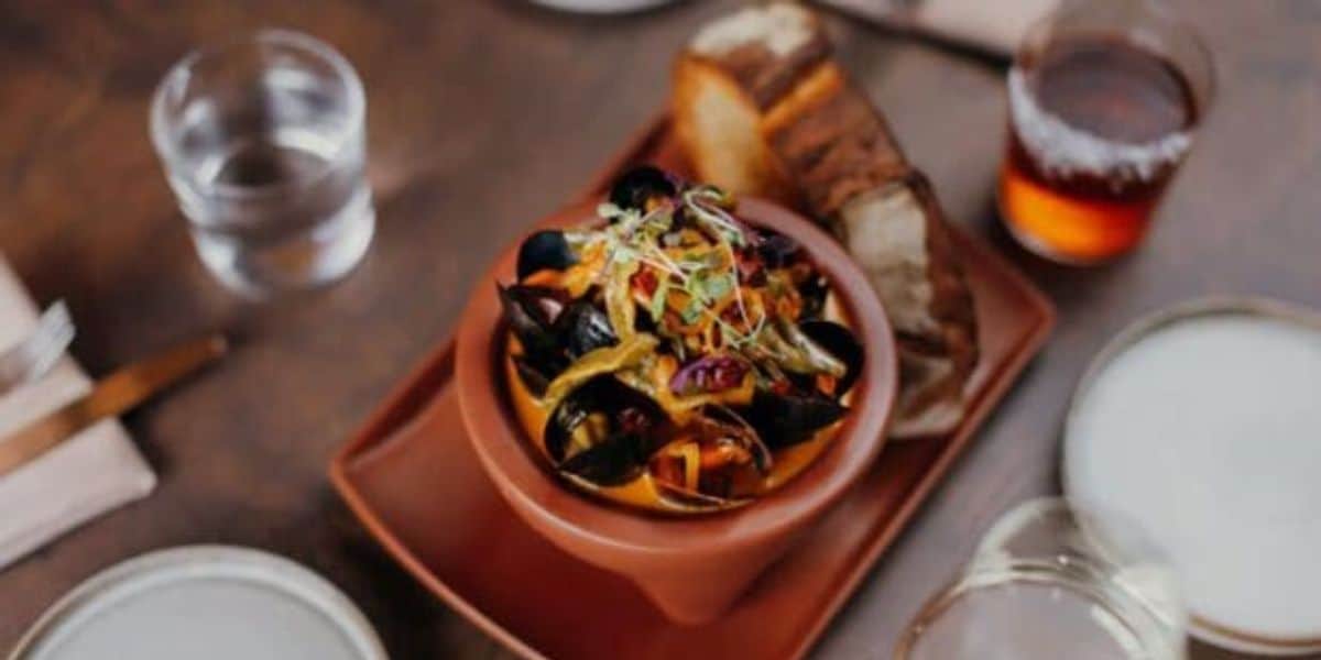 A bowl of mussels with vegetables and herbs sits on a wooden table, reminiscent of the vibrant flavors found at Downtown Disney restaurants. Accompanied by slices of bread and surrounded by glasses of water and an amber-colored beverage, it promises a delightful dining experience.