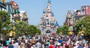 A bustling crowd fills the main street leading to a fairytale castle in Disneyland Paris, with pink walls and blue spires. The street is lined with green trees and colorful buildings under a clear, sunny sky.