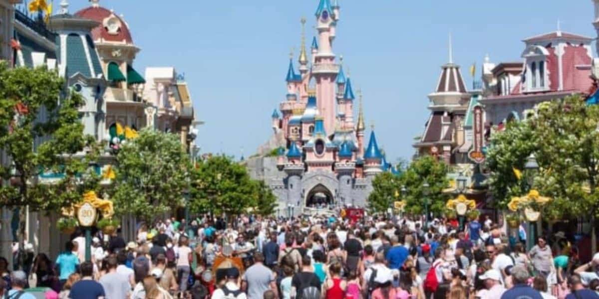 A bustling crowd fills the main street leading to a fairytale castle in Disneyland Paris, with pink walls and blue spires. The street is lined with green trees and colorful buildings under a clear, sunny sky.