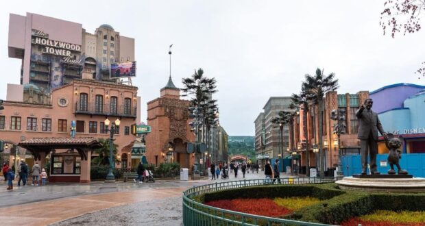 In this Disney Park scene, a statue of a man holding a mouse's hand graces the foreground. Surrounding buildings, including "The Hollywood Tower Hotel," showcase art deco architecture as people stroll along the wet pathways under a cloudy sky.