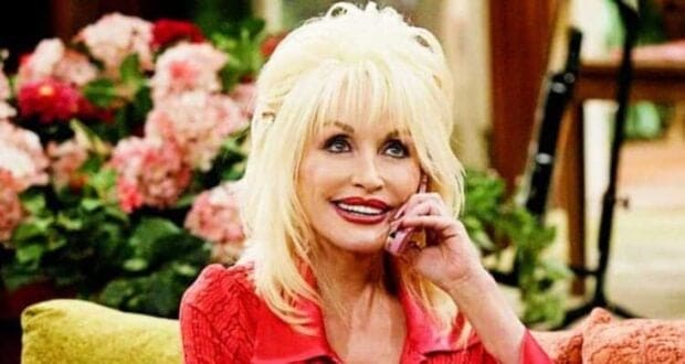 A woman with blonde hair styled with bangs and voluminous curls, wearing a red blouse, sits on a sofa with a phone, surrounded by pink floral arrangements.