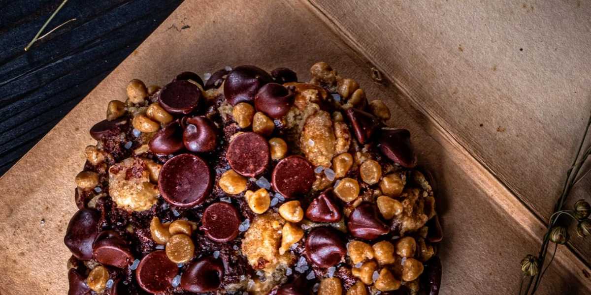 A close-up of a richly textured Gideons cookie topped with an assortment of chocolate chips and caramel pieces. The cookie is displayed on a wooden surface, with a dark background that subtly shows some dried twigs.