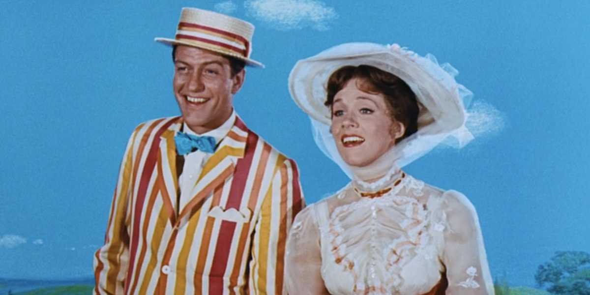 A smiling man and woman stand together against a blue sky with white clouds. The man wears a colorful striped suit, a bow tie, and a straw boater hat. The woman is dressed in a high-neck white dress with lace details and a wide-brimmed hat.