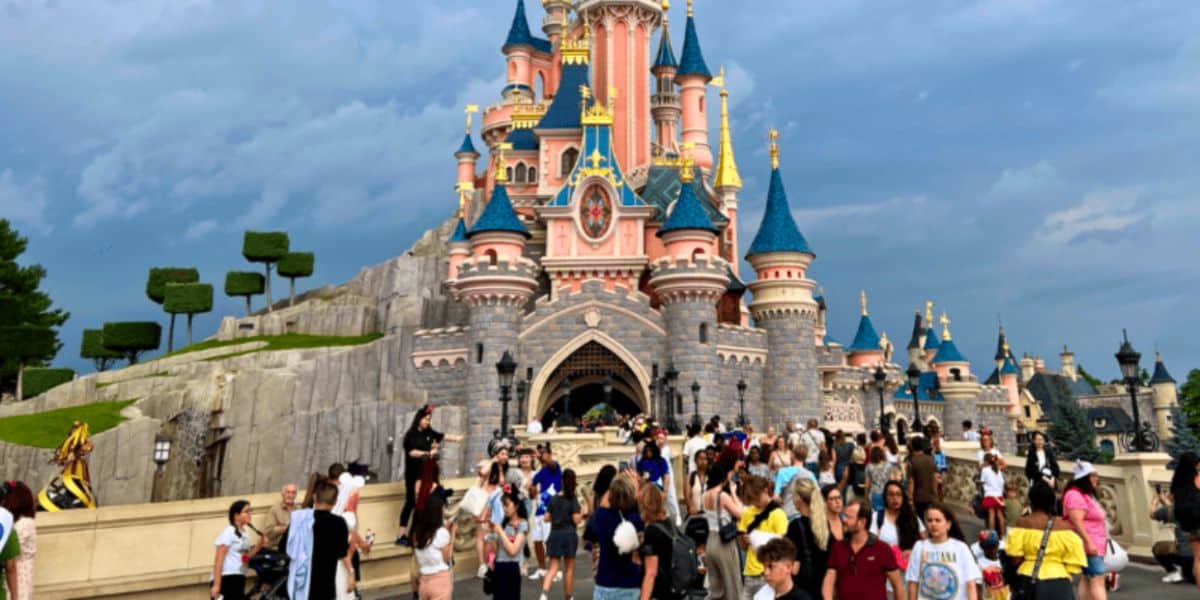 A bustling scene of visitors at Disneyland Paris in front of the iconic Sleeping Beauty Castle, with its pink and blue spires under a cloudy sky.