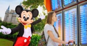 A person stands in front of a flight information board, looking up at the screen. To the left, an image of Mickey Mouse at a Disney theme park with a castle in the background adds a touch of magic to the scene.