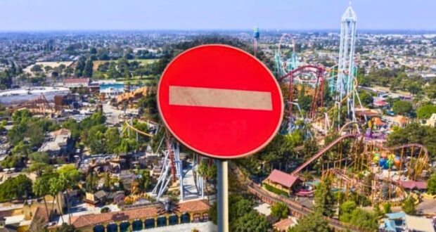 An aerial view of a theme park with various roller coasters and attractions, partially obscured by a large red and white "no entry" road sign in the foreground. The surrounding area includes trees and buildings.