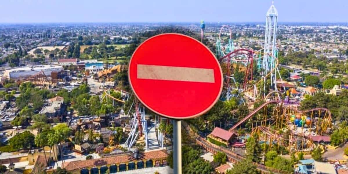 An aerial view of a theme park with various roller coasters and attractions, partially obscured by a large red and white "no entry" road sign in the foreground. The surrounding area includes trees and buildings.