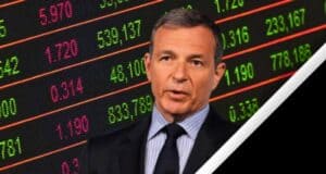 A man in a suit and striped tie speaks in front of a digital stock market display board showing various numbers and figures in red and green, including Disney Stock. The image has a diagonal white and black border on the right side.